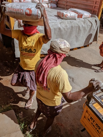 Two workers are carrying heavy bags of construction material, possibly cement, on their heads. They are wearing yellow shirts and have their faces covered with red cloths, likely for protection from dust or the sun. The scene takes place near a truck loaded with similar bags, indicating manual labor and transportation of goods.