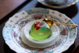 A decorative dessert is placed on an ornate, patterned plate with Asian-inspired motifs. The dessert is a translucent jelly with a green base and red and pink accents on top. A gold spoon rests next to the jelly, highlighting the luxurious presentation.