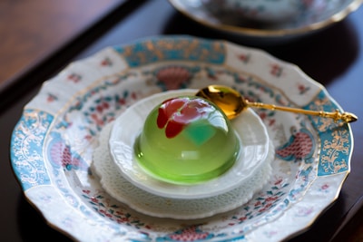 A decorative dessert is placed on an ornate, patterned plate with Asian-inspired motifs. The dessert is a translucent jelly with a green base and red and pink accents on top. A gold spoon rests next to the jelly, highlighting the luxurious presentation.
