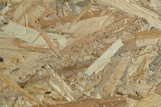 Close-up view of oriented strand board featuring a mix of wood chips and flakes pressed together. The texture is rough and uneven, showcasing various shades of beige and brown.