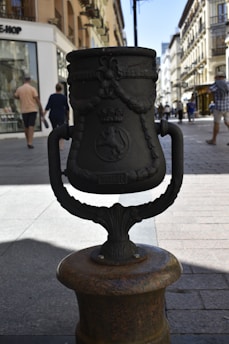 A large, ornate metal structure, possibly a decorative urban element, stands prominently in the foreground. It features intricate designs, including floral patterns, a crown, and an emblem. Behind it, people walk along a busy street lined with shops and historic buildings.