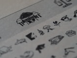 A series of small, detailed black and white illustrations on a textured paper. The images include various objects and symbols, such as a weather vane with a rooster, a tree, and scales, accompanied by other abstract and recognizable shapes. The paper has a grayish tone, and the illustrations are sharp against the slightly blurred background.