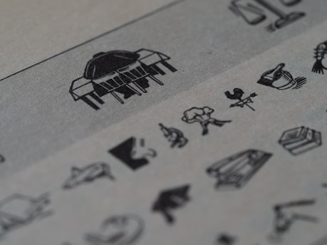 A series of small, detailed black and white illustrations on a textured paper. The images include various objects and symbols, such as a weather vane with a rooster, a tree, and scales, accompanied by other abstract and recognizable shapes. The paper has a grayish tone, and the illustrations are sharp against the slightly blurred background.