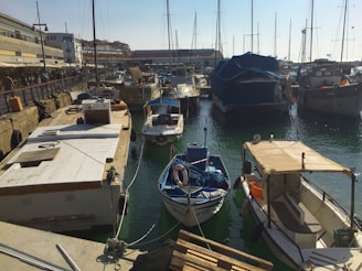 Boats are moored in a bustling harbor area, with wooden piers and a variety of vessels, including yachts and smaller boats. The water is a deep green, reflecting the light from a clear sky. Buildings line the harbor, with people walking along the waterfront, and tall masts create a dense, vertical pattern against the horizon.