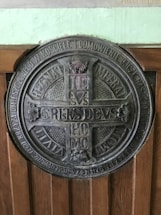 A large circular metal plaque featuring detailed engravings and inscriptions in Latin. The center of the plaque contains a cross-like design with letters arranged symmetrically. Surrounding the central motif, additional text encircles the entire piece, with decorative elements such as floral patterns. The plaque is mounted on a wooden surface with visible grain.