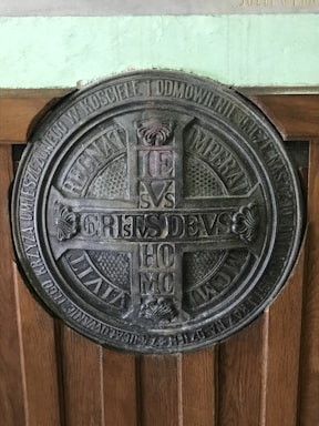 A large circular metal plaque featuring detailed engravings and inscriptions in Latin. The center of the plaque contains a cross-like design with letters arranged symmetrically. Surrounding the central motif, additional text encircles the entire piece, with decorative elements such as floral patterns. The plaque is mounted on a wooden surface with visible grain.