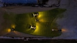 Three tractors with bright headlights illuminate a field in a dark, industrial or agricultural setting. The lights create distinctive yellow-green patches on the ground, and the machinery appears to be active in a coordinated manner.