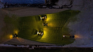 Three tractors with bright headlights illuminate a field in a dark, industrial or agricultural setting. The lights create distinctive yellow-green patches on the ground, and the machinery appears to be active in a coordinated manner.
