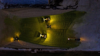 Three tractors with bright headlights illuminate a field in a dark, industrial or agricultural setting. The lights create distinctive yellow-green patches on the ground, and the machinery appears to be active in a coordinated manner.