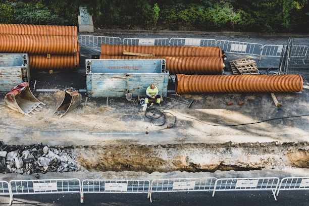 A construction site with large orange pipes and metal barriers. A person in a high-visibility vest is sitting near a trench, surrounded by construction equipment. Excavators and other machinery parts are present, and the ground is partially dug up with visible rubble.