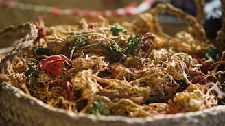 A woven basket filled with assorted dried flowers and herbs, featuring intertwined strands of various shapes and sizes. The colors include earthy tones and some pops of red and green, creating a rustic and natural appearance.