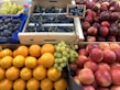 A fruit market display featuring various fruits neatly arranged in crates and baskets. There are bright orange oranges, shiny red apples, deep purple figs, and clusters of green and dark purple grapes. The fruits have a fresh and vibrant appearance, with a sign indicating the price of the grapes.