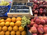 A fruit market display featuring various fruits neatly arranged in crates and baskets. There are bright orange oranges, shiny red apples, deep purple figs, and clusters of green and dark purple grapes. The fruits have a fresh and vibrant appearance, with a sign indicating the price of the grapes.