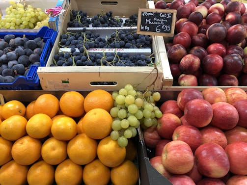 A fruit market display featuring various fruits neatly arranged in crates and baskets. There are bright orange oranges, shiny red apples, deep purple figs, and clusters of green and dark purple grapes. The fruits have a fresh and vibrant appearance, with a sign indicating the price of the grapes.