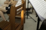 A room contains a wooden chair with some Chinese characters on the backrest. A person is seated, partially visible, wearing dark shorts and holding a violin bow. A music stand with sheet music is positioned in the foreground.