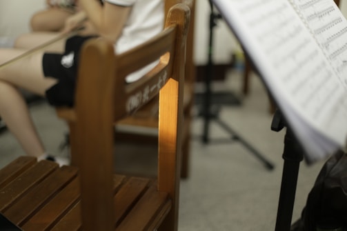 A room contains a wooden chair with some Chinese characters on the backrest. A person is seated, partially visible, wearing dark shorts and holding a violin bow. A music stand with sheet music is positioned in the foreground.