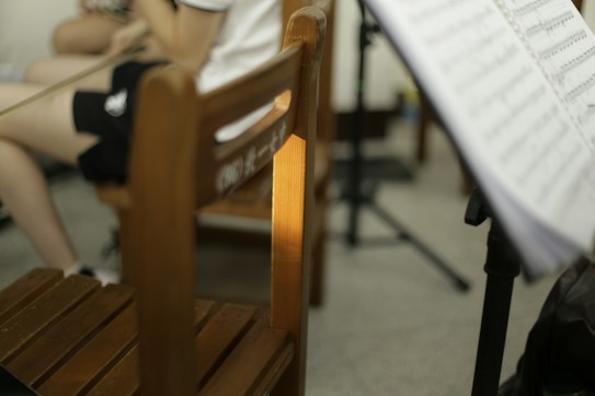 A room contains a wooden chair with some Chinese characters on the backrest. A person is seated, partially visible, wearing dark shorts and holding a violin bow. A music stand with sheet music is positioned in the foreground.