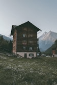A rustic, multi-story wooden building stands amidst a picturesque mountainous landscape. The structure is surrounded by lush green grass and small wildflowers. Majestic mountains rise in the background under a clear, dusky sky, adding a serene and remote feel to the scene.