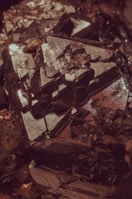An intricate cluster of dark, metallic crystals reflecting light with sharp and geometric surfaces. The shapes are angular, with some parts appearing as if they are jutting out from the main body of the mineral. The overall texture is rough, with various facets catching subtle highlights.