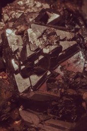 An intricate cluster of dark, metallic crystals reflecting light with sharp and geometric surfaces. The shapes are angular, with some parts appearing as if they are jutting out from the main body of the mineral. The overall texture is rough, with various facets catching subtle highlights.
