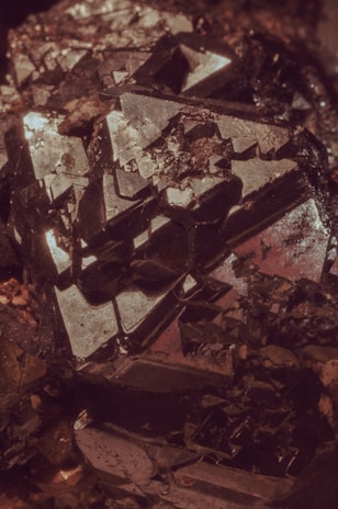 An intricate cluster of dark, metallic crystals reflecting light with sharp and geometric surfaces. The shapes are angular, with some parts appearing as if they are jutting out from the main body of the mineral. The overall texture is rough, with various facets catching subtle highlights.