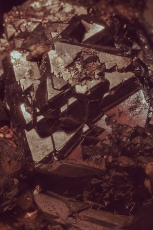 An intricate cluster of dark, metallic crystals reflecting light with sharp and geometric surfaces. The shapes are angular, with some parts appearing as if they are jutting out from the main body of the mineral. The overall texture is rough, with various facets catching subtle highlights.
