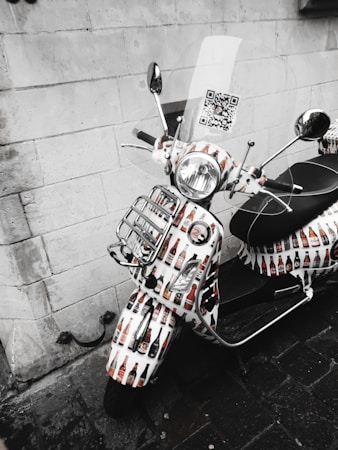 A scooter decorated with images of beer bottles is parked against a stone wall. The front is adorned with various bottle designs, and a QR code is visible on the windshield. The scene is devoid of color except for the decorated parts of the scooter, lending a striking effect against the grayscale background.