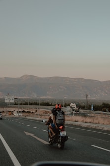 A motorcycle with two riders travels on a highway surrounded by mountains and greenery. The rider in front wears a white shirt while the one in back has a black helmet and a gray backpack. The road is clear with a few vehicles, and there is a sign with text on the left. The sky is clear, and it's likely taken during late afternoon or early evening.