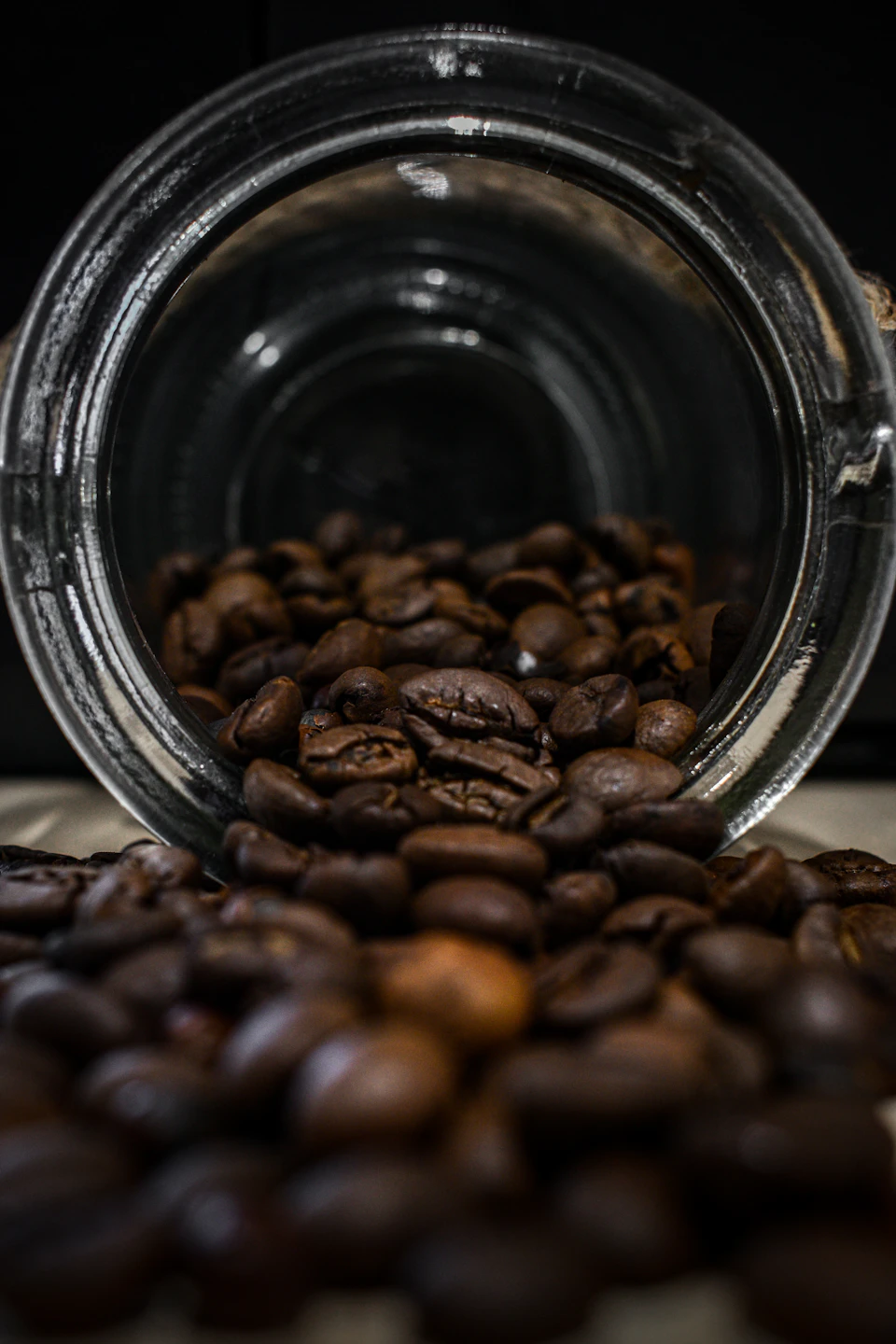 The image shows a glass jar filled with coffee beans spilling out onto a white surface, creating an interesting visual effect. The coffee beans are dark brown and scattered across the surface, some overlapping each other while others remain separate. The background is blurred, drawing focus to the jar of coffee beans as the main subject in the scene.
