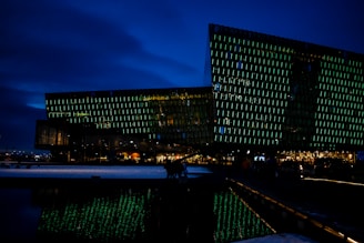 A modern, angular building with a glass facade illuminated by green lights, set against a dark blue night sky. The building's unique design reflects in the water in the foreground. Warm, interior lights glow from inside, suggesting activity within.