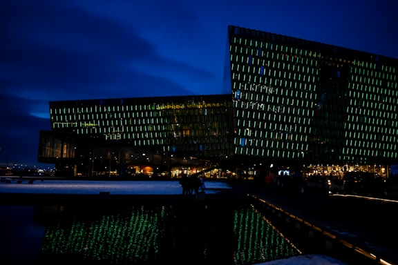 A modern, angular building with a glass facade illuminated by green lights, set against a dark blue night sky. The building's unique design reflects in the water in the foreground. Warm, interior lights glow from inside, suggesting activity within.