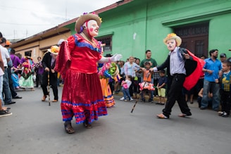 A group of people is participating in a colorful street parade. Participants are wearing vibrant costumes and masks, with one person dressed in a red and blue dress and another in a black outfit with a cape and straw hat. The crowd of onlookers, including adults and children, stands along the side of the street, watching the procession.
