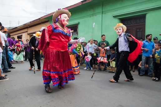 A group of people is participating in a colorful street parade. Participants are wearing vibrant costumes and masks, with one person dressed in a red and blue dress and another in a black outfit with a cape and straw hat. The crowd of onlookers, including adults and children, stands along the side of the street, watching the procession.