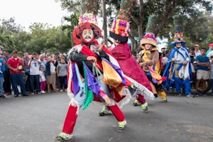 A vibrant street performance featuring dancers in colorful, elaborate costumes. The dancers wear lavishly decorated hats and masks with exaggerated features, adding to the festive atmosphere. They are moving energetically on a paved street with a backdrop of a crowd of onlookers and trees.