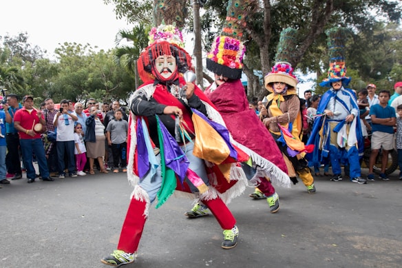 A vibrant street performance featuring dancers in colorful, elaborate costumes. The dancers wear lavishly decorated hats and masks with exaggerated features, adding to the festive atmosphere. They are moving energetically on a paved street with a backdrop of a crowd of onlookers and trees.