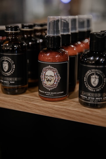A display of grooming products featuring several bottles of aftershave lotion with black and brown colored labels. The bottles are arranged on a wooden shelf with a soft focus in the background, highlighting the central product which has an intricate label design featuring a barbershop emblem.