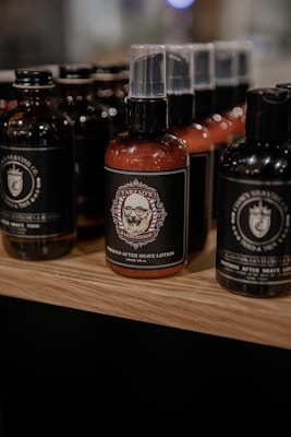 A display of grooming products featuring several bottles of aftershave lotion with black and brown colored labels. The bottles are arranged on a wooden shelf with a soft focus in the background, highlighting the central product which has an intricate label design featuring a barbershop emblem.