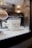 A white shaving mug labeled 'Parker Safety Razor' is on display, containing several packaged razor blades. Nearby, a shaving brush with a wooden handle and soft bristles is partially visible. The setting appears to be a well-lit, elegant bathroom or barbershop with a reflective surface.