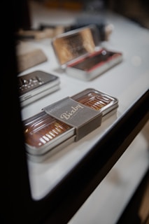A sleek metal box displayed on a glass surface. It has a transparent lid revealing organized contents inside and features a stylish label. Several similar tins are blurred in the background.