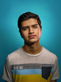 A young person with short dark hair is wearing a light-colored t-shirt with the text 'BLUE INC' and a multicolored stripe design, standing against a bright blue background. The mood appears neutral and contemplative.