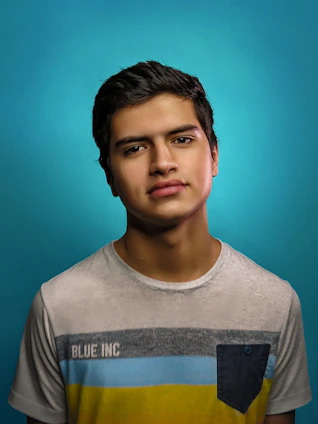 A young person with short dark hair is wearing a light-colored t-shirt with the text 'BLUE INC' and a multicolored stripe design, standing against a bright blue background. The mood appears neutral and contemplative.