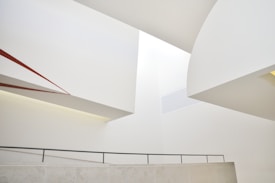 An abstract architectural composition featuring intersecting geometric forms and clean lines. The walls and ceiling are predominantly white, with a touch of red accent on one of the diagonal elements. The space conveys a sense of modern minimalism through its smooth surfaces and muted tones.
