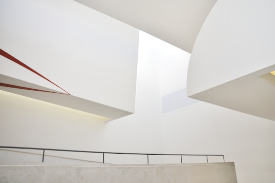 An abstract architectural composition featuring intersecting geometric forms and clean lines. The walls and ceiling are predominantly white, with a touch of red accent on one of the diagonal elements. The space conveys a sense of modern minimalism through its smooth surfaces and muted tones.