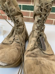 A pair of worn-out, dusty combat boots with visible scuff marks and frayed edges are shown up close. The boots have long laces, some of which are loosely tied, and military dog tags are placed on top of one of the boots. The background features a brick wall and a strip of green paint.
