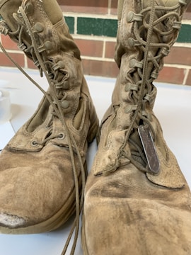 A pair of worn-out, dusty combat boots with visible scuff marks and frayed edges are shown up close. The boots have long laces, some of which are loosely tied, and military dog tags are placed on top of one of the boots. The background features a brick wall and a strip of green paint.