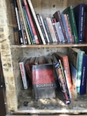 A collection of books arranged on a wooden bookshelf, with some books standing upright and others leaning or stacked. The shelves are lined with vintage-style newspaper clippings. A prominent book titled 'Bourdieu: Journalistic Field' is leaning forward, drawing attention with its distinct red and black cover.