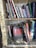 A collection of books arranged on a wooden bookshelf, with some books standing upright and others leaning or stacked. The shelves are lined with vintage-style newspaper clippings. A prominent book titled 'Bourdieu: Journalistic Field' is leaning forward, drawing attention with its distinct red and black cover.