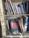 A collection of books arranged on a wooden bookshelf, with some books standing upright and others leaning or stacked. The shelves are lined with vintage-style newspaper clippings. A prominent book titled 'Bourdieu: Journalistic Field' is leaning forward, drawing attention with its distinct red and black cover.