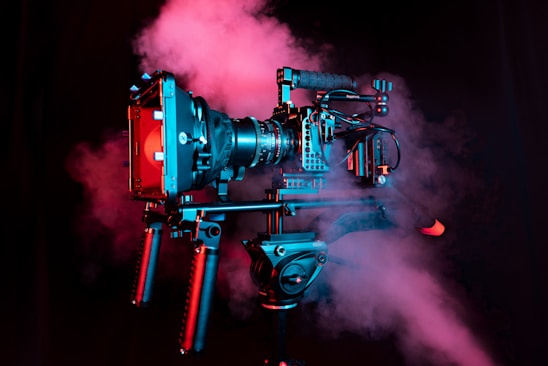 A professional video camera setup on a tripod is enveloped in pink and blue haze. The camera is adorned with various attachments and handles, indicating a high-end, cinematic rig used in professional filming.
