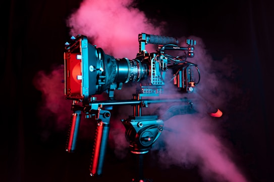 A professional video camera setup on a tripod is enveloped in pink and blue haze. The camera is adorned with various attachments and handles, indicating a high-end, cinematic rig used in professional filming.